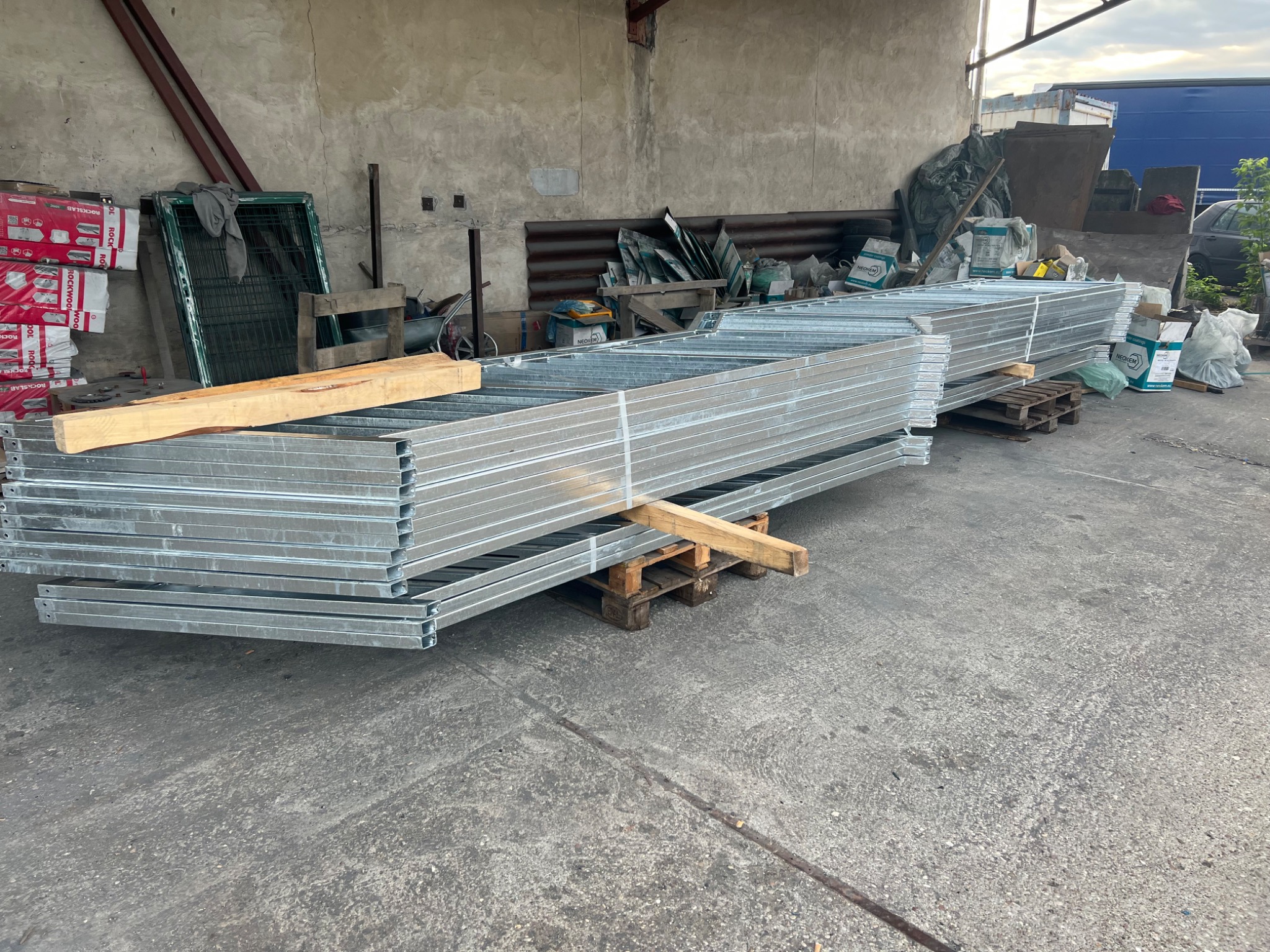 Stos ocynkowanych, metalowych ram na paletach, prawdopodobnie elementów ogrodzenia lub bramy, składowanych w magazynie lub na placu budowy.