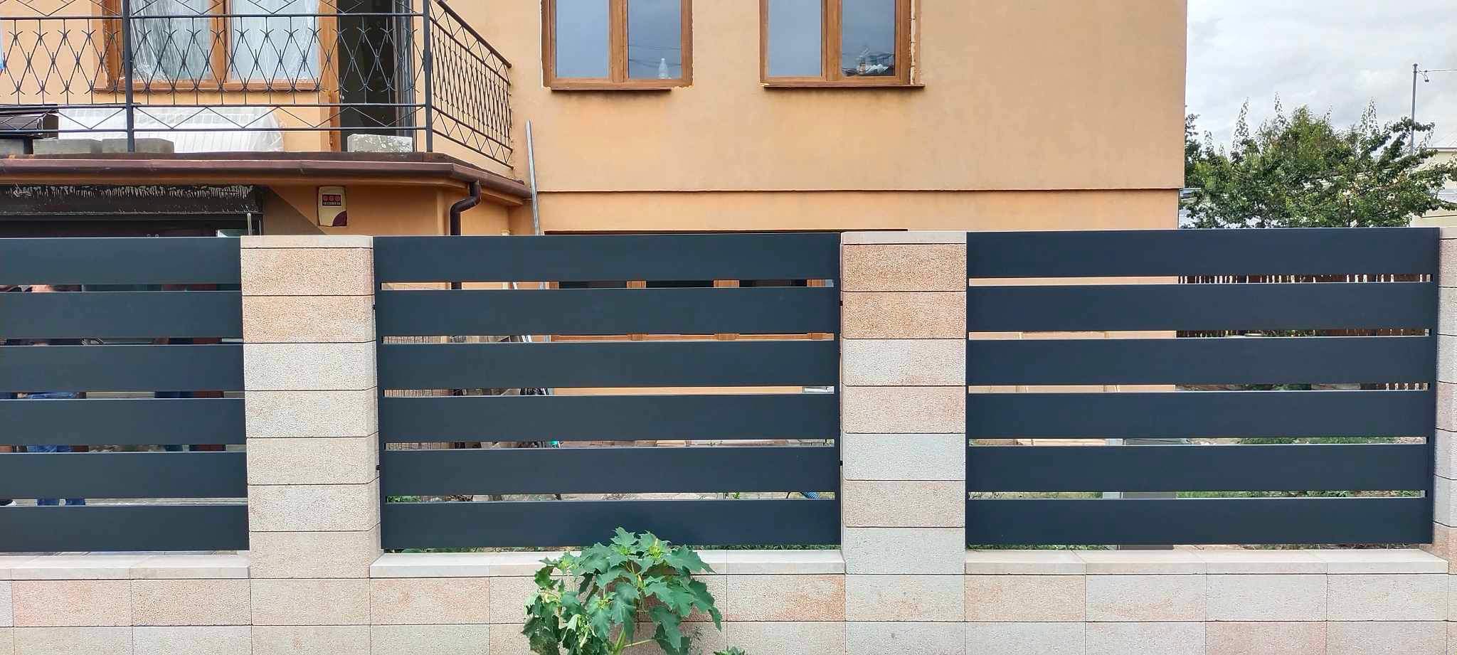 Nowoczesne ogrodzenie poziome z metalowymi przęsłami i kamiennymi słupkami w Białymstoku, widok z poziomu gruntu, z roślinnością u podstawy.