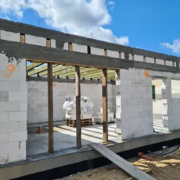 Wznoszenie ścian parterowego domu z betonu komórkowego, widoczne drewniane belki stropowe i elementy szalunku, pomarańczowe oznaczenia na ścianach, białe bloczki na palecie wewnątrz budynku.