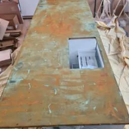 Blat roboczy z efektem patyny, z wyciętym otworem na szafkę, zabezpieczony folią malarską, w tle kartony i kozły malarskie.