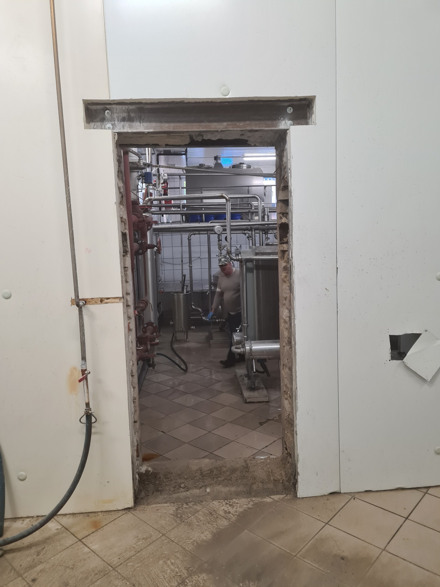 Prace remontowe w pomieszczeniu przemysłowym: Wykucie otworu drzwiowego w ścianie z płyt warstwowych, widoczne instalacje i pracownik w tle.