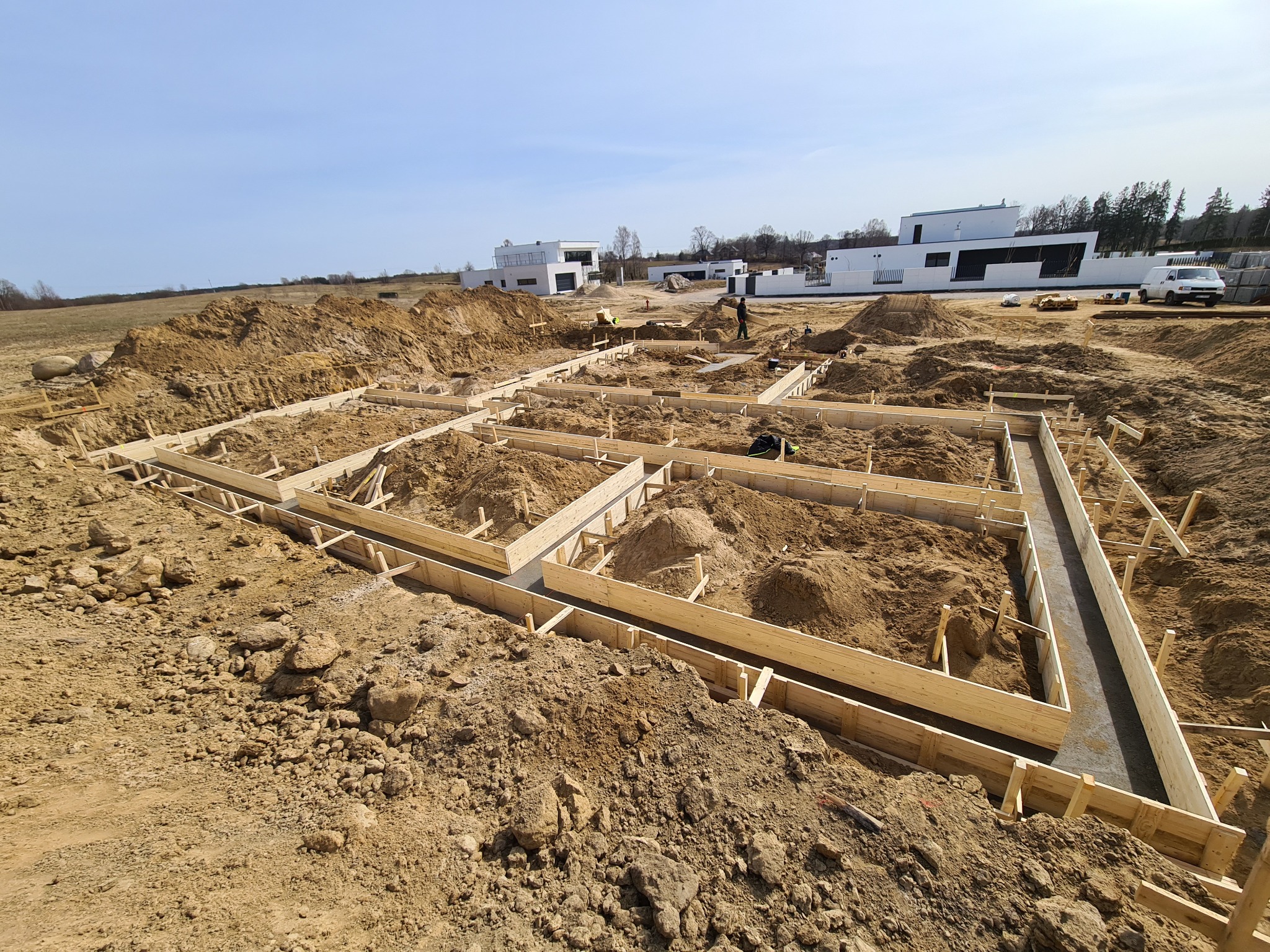 Przygotowanie fundamentów pod budowę domu: drewniane szalunki zalane betonem na placu budowy z widocznymi pracownikami i nowoczesnymi domami w tle.