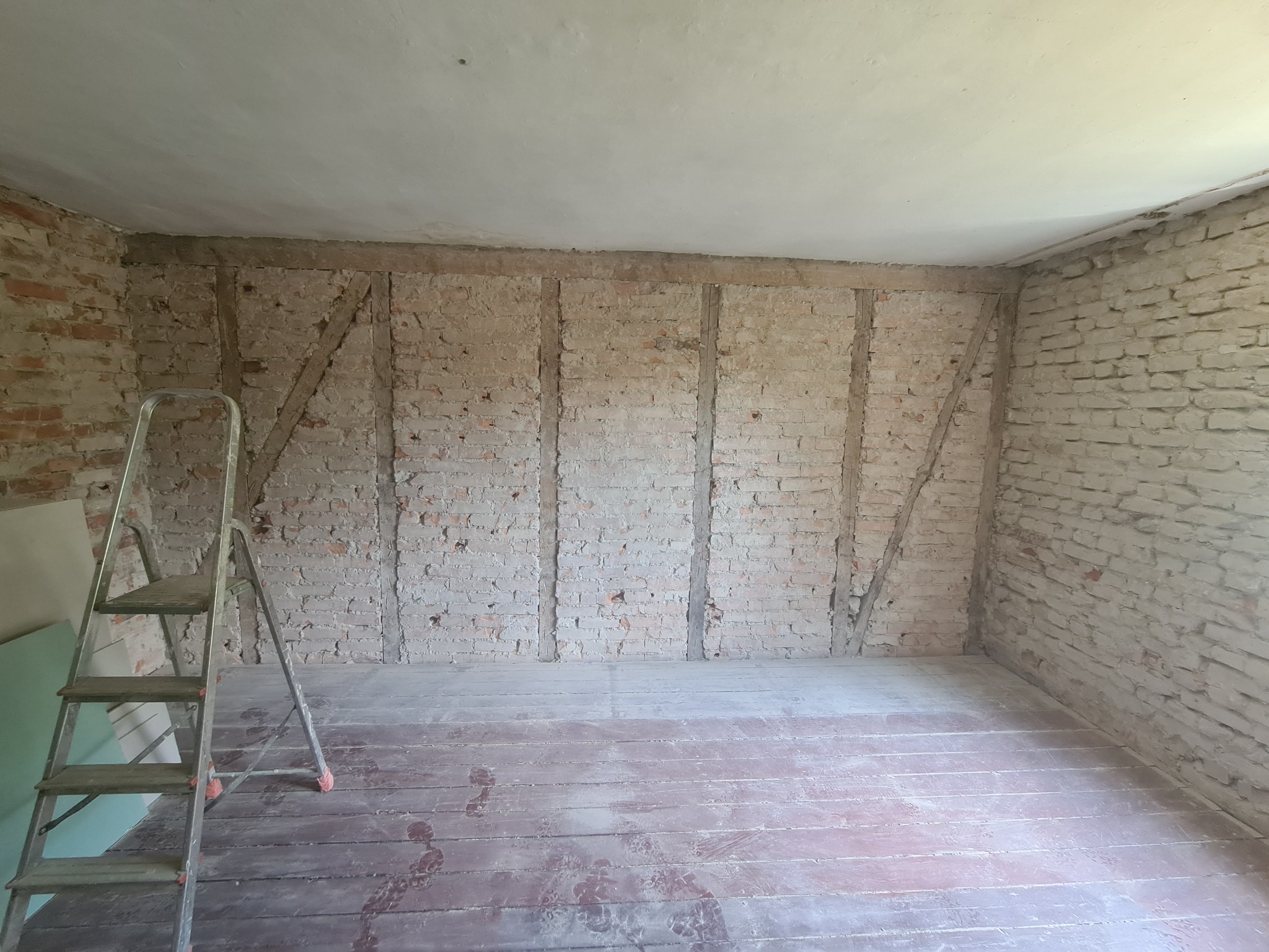 Pokój w trakcie remontu: odsłonięta cegła na ścianach, drewniane elementy konstrukcyjne, stara drewniana podłoga, drabina oparta o ścianę, płyty gipsowe w rogu.