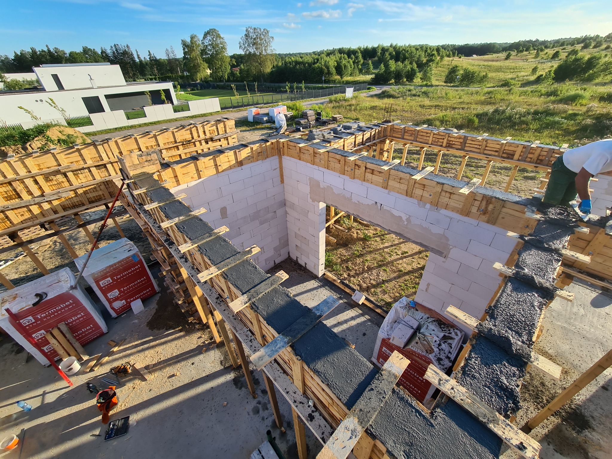 Budowa domu z betonu komórkowego: widok z góry na wznoszone ściany i zalewane betonem szalunki, w tle nowoczesny dom jednorodzinny i zielony krajobraz.