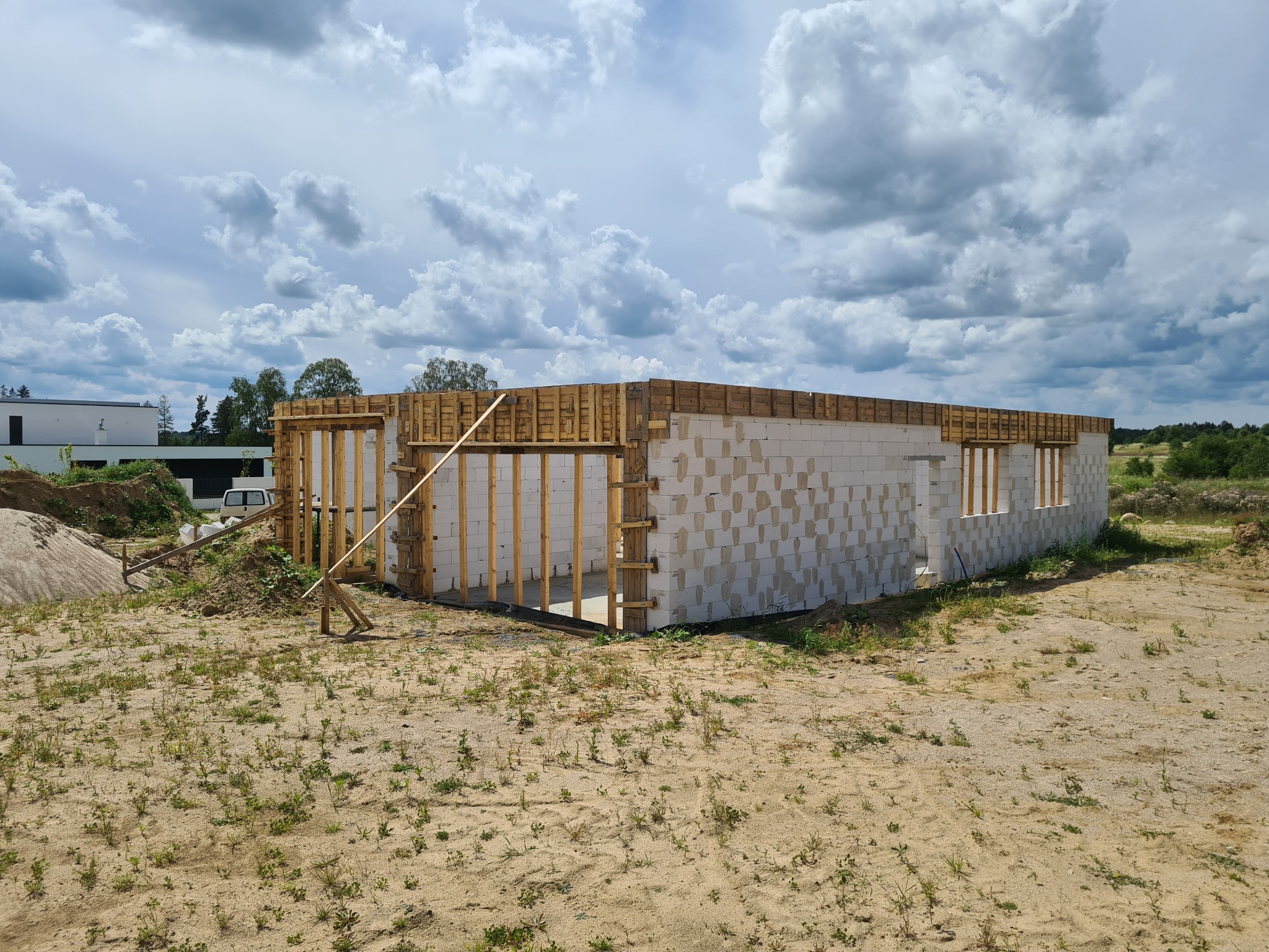 Surowy stan domu z białych bloczków, widoczne drewniane szalunki okienne i wieńce, na piaszczystym terenie z rzadką roślinnością, pod pochmurnym niebem.