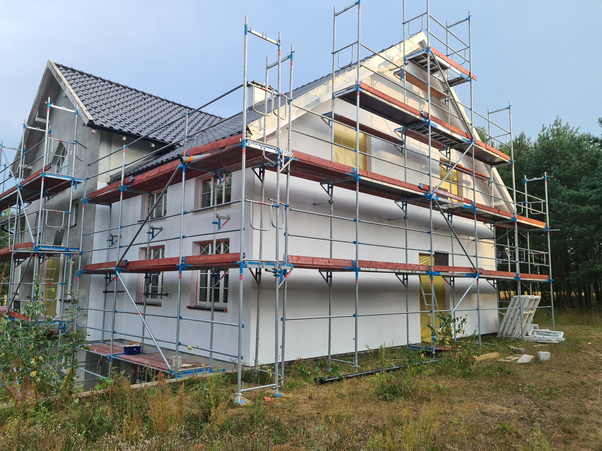 Dom murowany w trakcie ocieplania elewacji, widoczne rusztowania, częściowo otynkowana ściana i fragmenty ocieplenia poddasza, nowe okna.