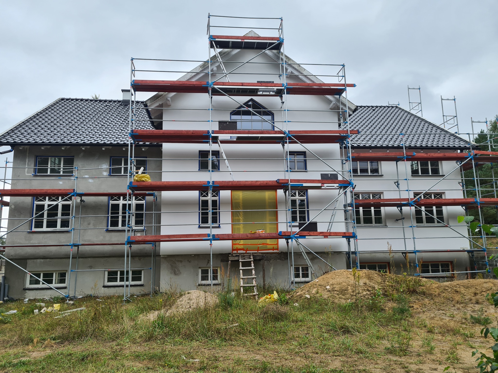 Dwukondygnacyjny dom murowany w trakcie prac elewacyjnych, otoczony rusztowaniami, z częściowo otynkowaną i pomalowaną fasadą, zabezpieczonymi oknami folią i stertą piasku na pierwszym planie.