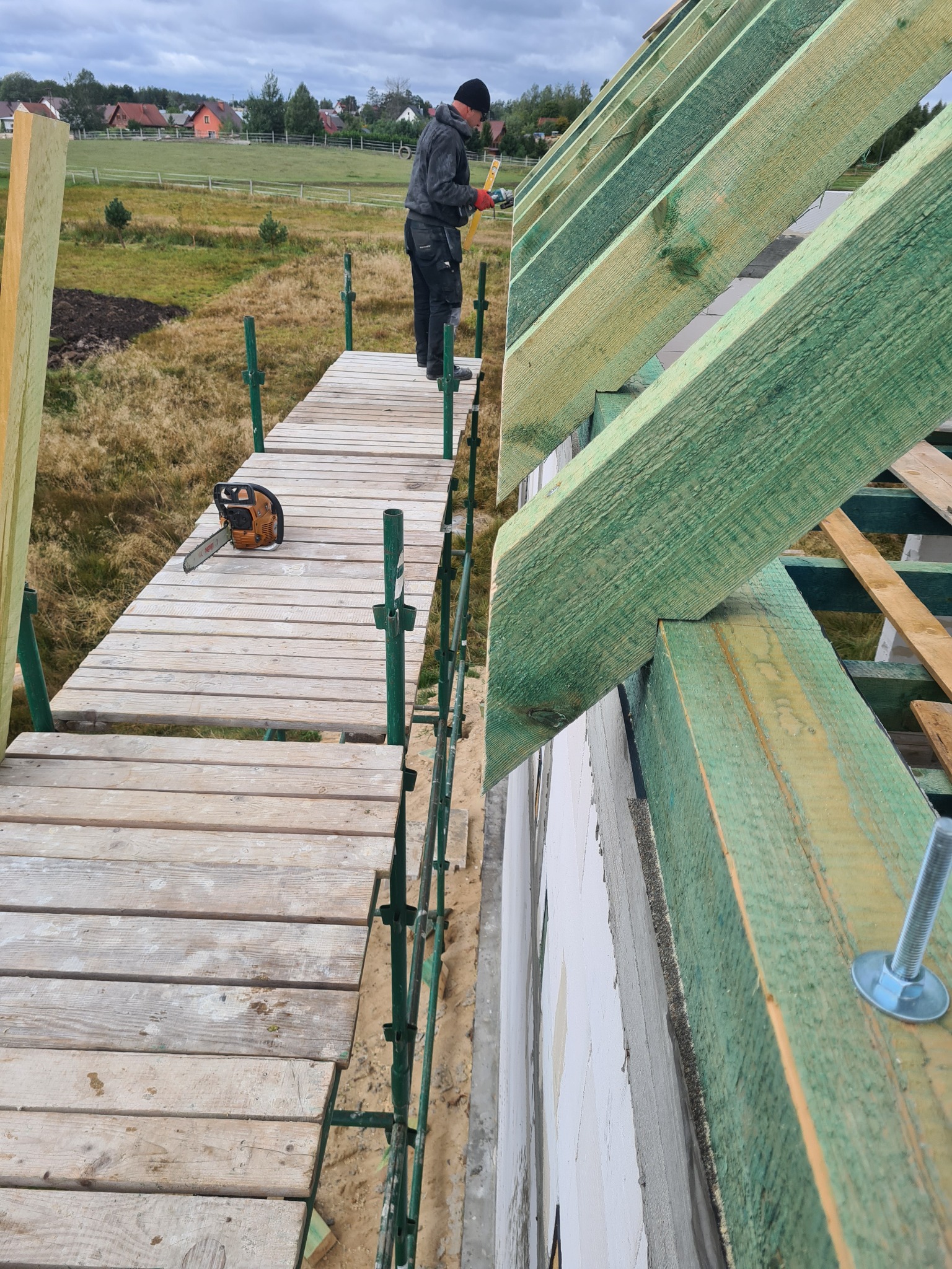 Budowa dachu: Widok z rusztowania na konstrukcję drewnianą zaimpregnowaną na zielono, mocowaną do ściany domu z betonu komórkowego. Na rusztowaniu leży piła spalinowa, a w tle widać krajobraz wiejski.