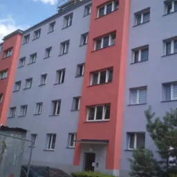 Odnowiony blok mieszkalny z szarą elewacją i pionowymi, pomarańczowymi akcentami, widoczny napis 'Głowackiego 27B' oraz fragment ogrodzenia budowy z napisem 'Teren Budowy Wstęp Zabroniony'.