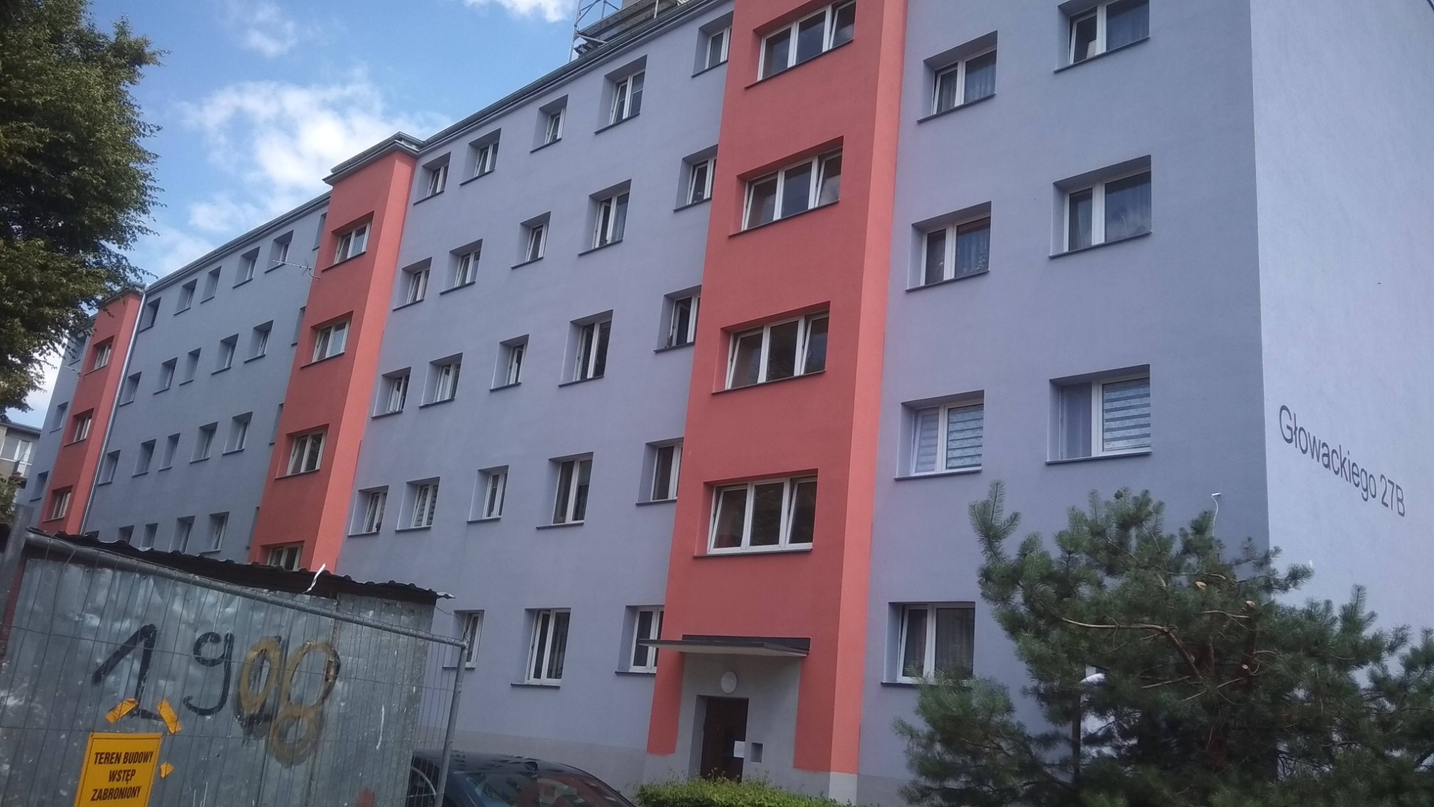 Odnowiony blok mieszkalny z szarą elewacją i pionowymi, pomarańczowymi akcentami, widoczny napis 'Głowackiego 27B' oraz fragment ogrodzenia budowy z napisem 'Teren Budowy Wstęp Zabroniony'.