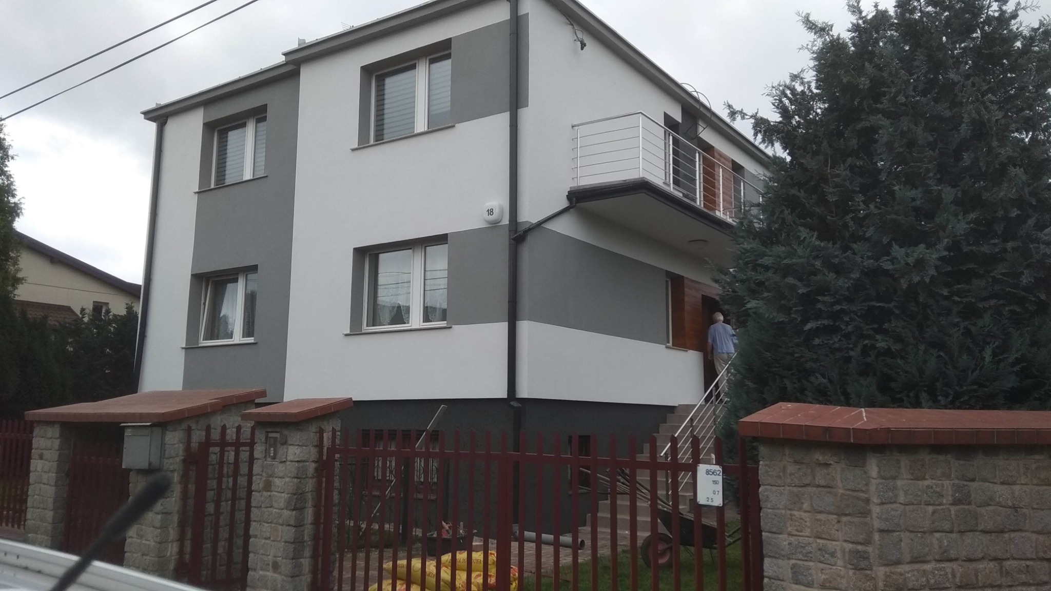 Dwukondygnacyjny dom z balkonem i elewacją w odcieniach szarości i bieli, widoczny zza czerwonego ogrodzenia i zieleni, z osobą wchodzącą po schodach.