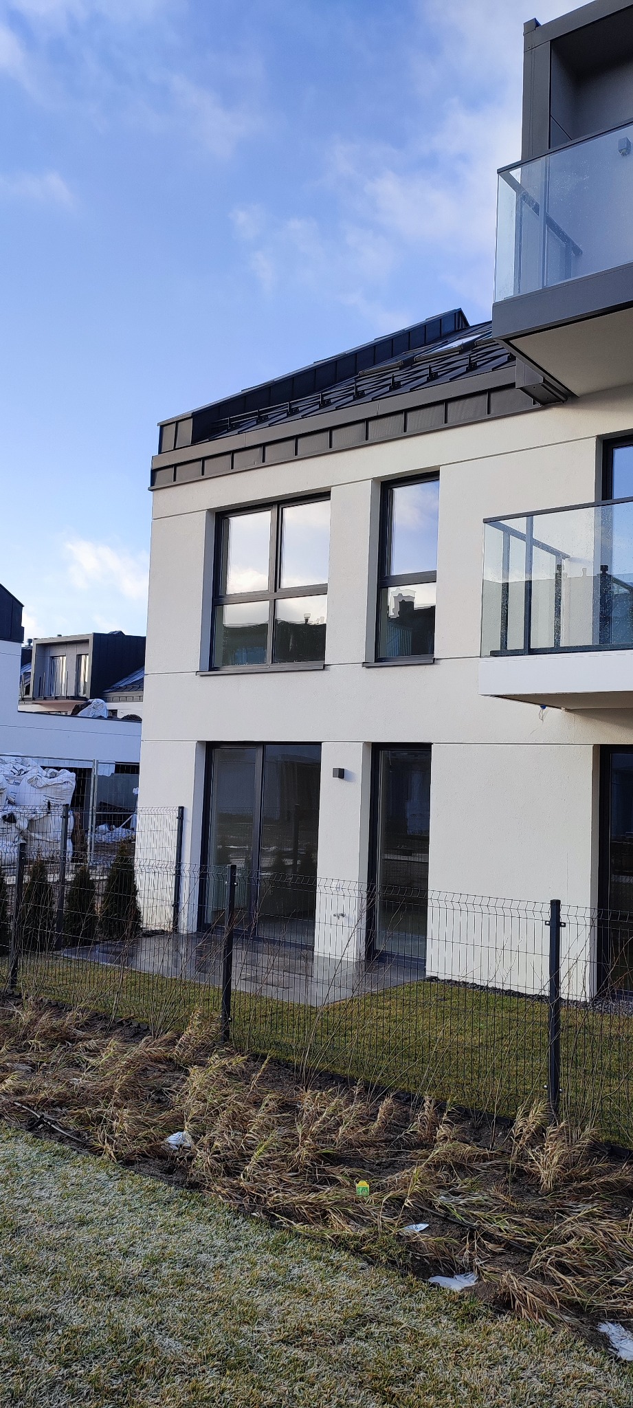 Nowoczesny budynek z jasną elewacją, ciemnymi ramami okiennymi i szklaną balustradą balkonu, otoczony metalowym ogrodzeniem i częściowo zamarzniętą trawą.