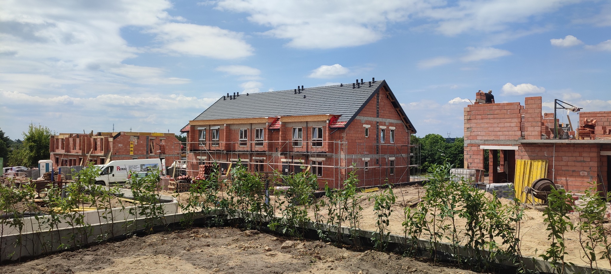 Budowa bliźniaczych domów z czerwonej cegły, z szarym dachem i rusztowaniami, w tle częściowo wykończone domy, błękitne niebo z chmurami i zielona roślinność.