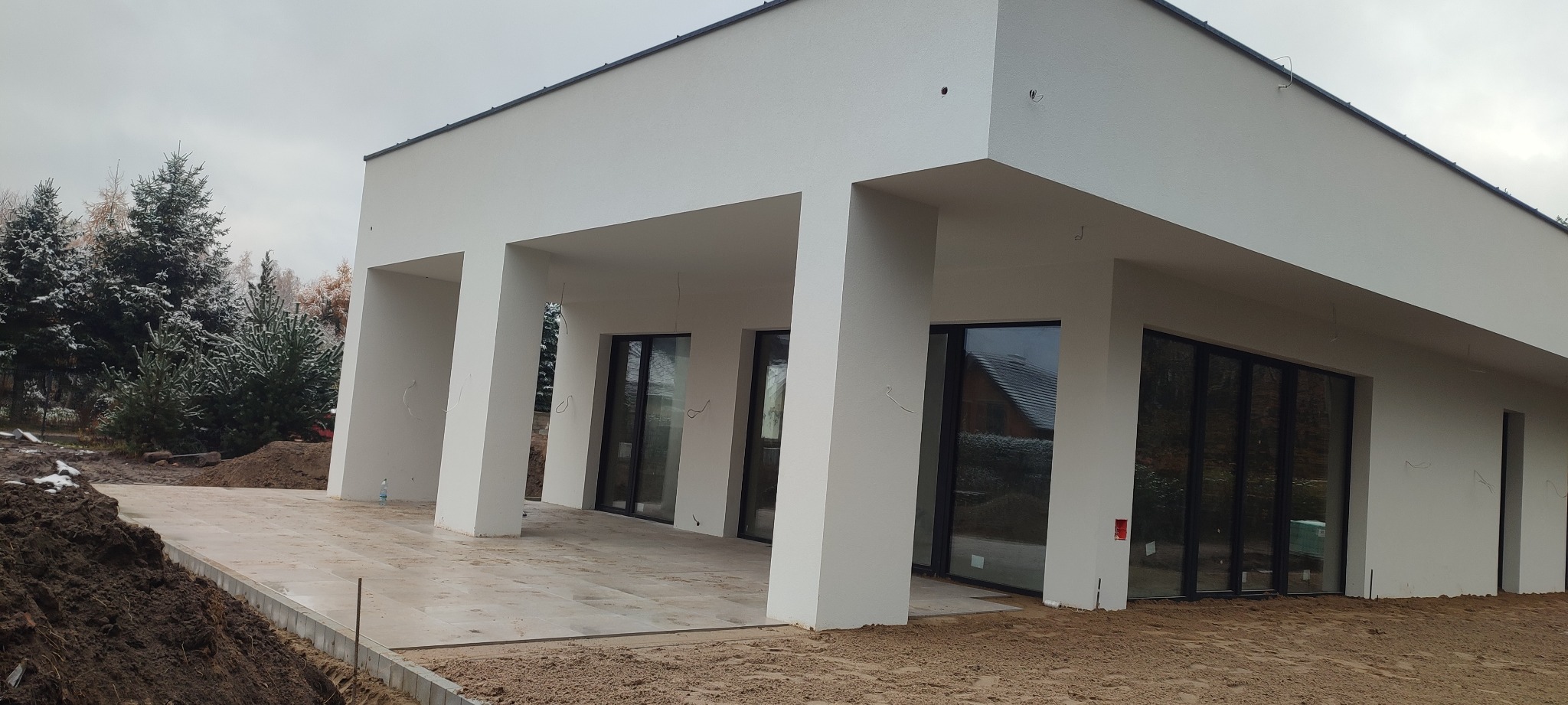 Nowoczesny dom z białymi ścianami i dużymi oknami, widok z zewnątrz, teren wokół w trakcie prac wykończeniowych, widoczny fragment tarasu i przygotowane podłoże.