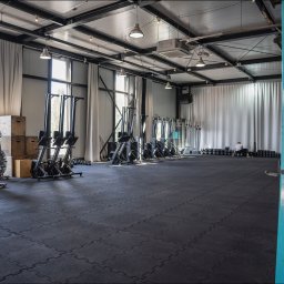 Nieruchome - Przestronna sala fitness z czarną gumową podłogą, rzędem ergometrów wioślarskich, workami bokserskimi, hantlami i stalową konstrukcją sufitu z oświetleniem punktowym.