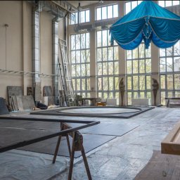 Nieruchome - Przestronne wnętrze hali produkcyjnej z dużymi oknami, widoczne elementy scenografii, drewniane konstrukcje i niebieska draperia pod sufitem.