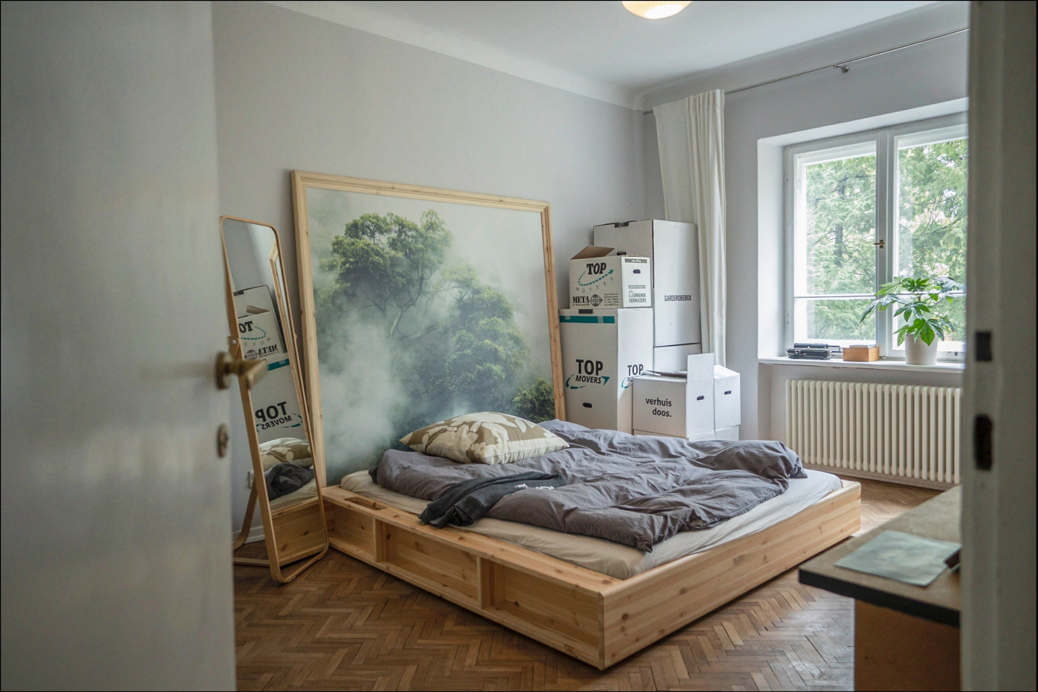 Jasne wnętrze sypialni z drewnianym łóżkiem platformowym, dużym obrazem z motywem leśnym, lustrem i kartonami po przeprowadzce, parkiet w jodełkę na podłodze.