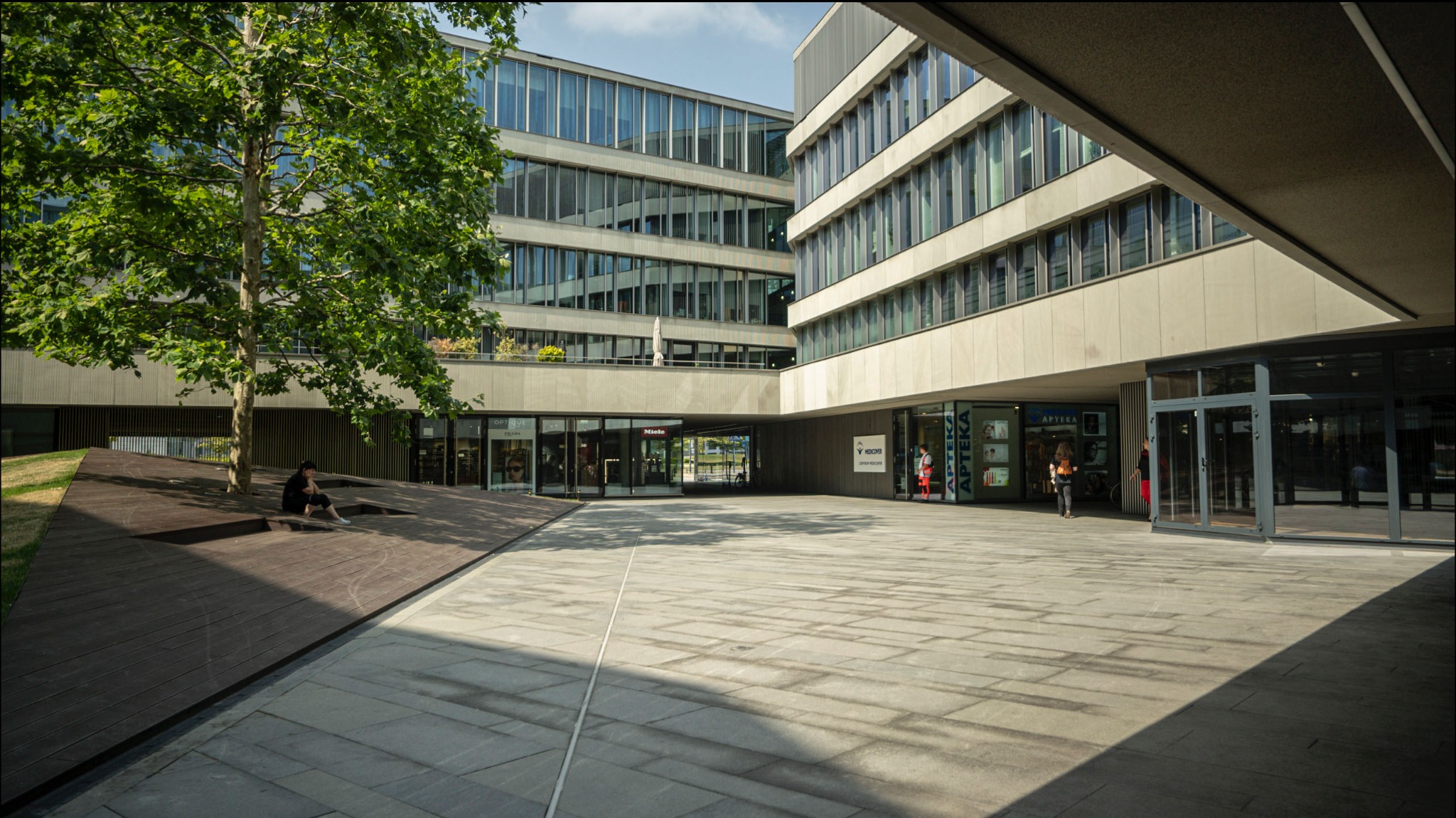 Kwadratowy dziedziniec biurowy z nowoczesną architekturą, drzewem i osobą siedzącą na drewnianym podeście. W tle widoczne witryny sklepowe.