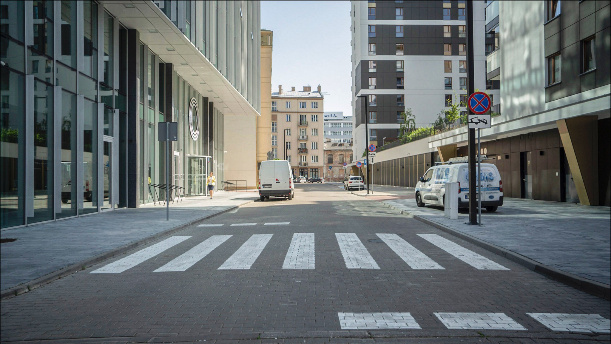 Ulica miejska z przejściem dla pieszych na pierwszym planie, w tle nowoczesne budynki biurowe i mieszkalne, zaparkowane busy dostawcze po obu stronach drogi.