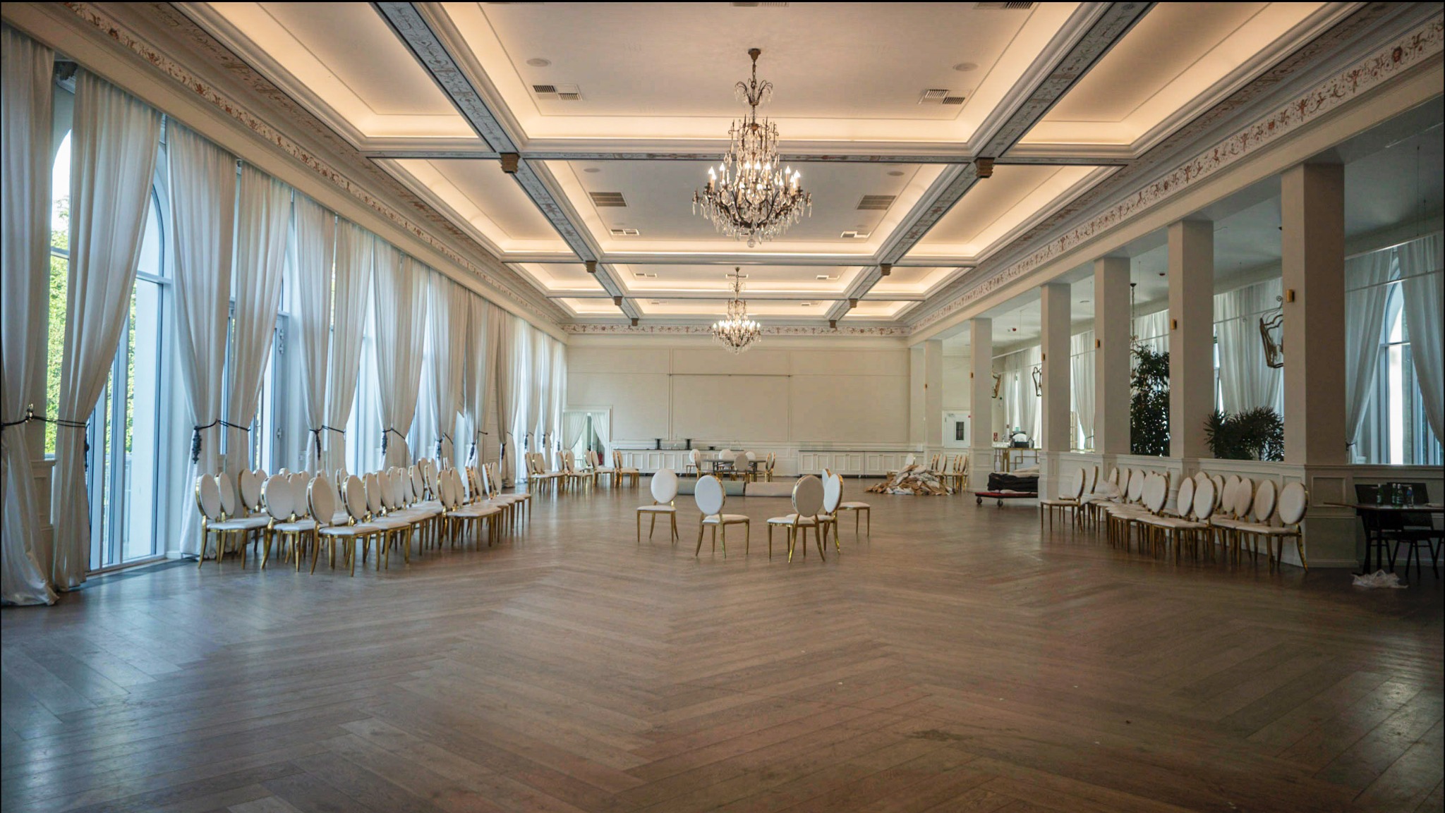 Przestronne wnętrze sali balowej z parkietem w jodełkę, wysokimi oknami z białymi zasłonami, kryształowymi żyrandolami i krzesłami ustawionymi w rzędach i okręgach.