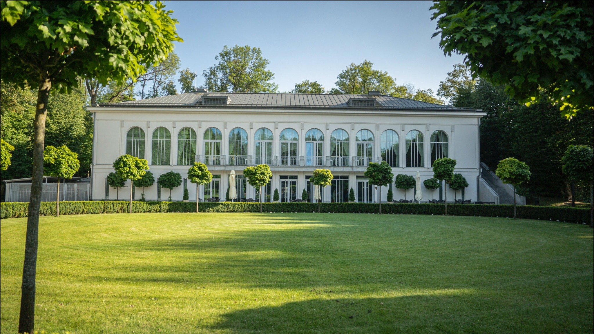 Elegancki, biały budynek z dużymi oknami łukowymi i balkonami, otoczony zadbanym trawnikiem i formowanymi drzewami.