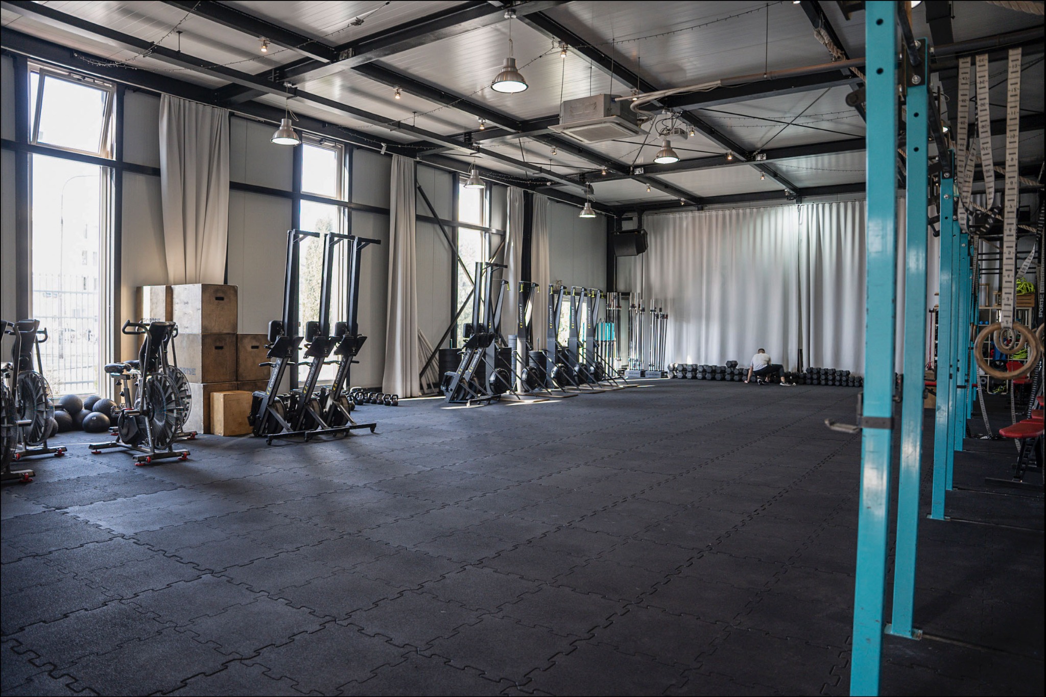 Przestronna sala fitness z czarną gumową podłogą, rzędem ergometrów wioślarskich, workami bokserskimi, hantlami i stalową konstrukcją sufitu z oświetleniem punktowym.