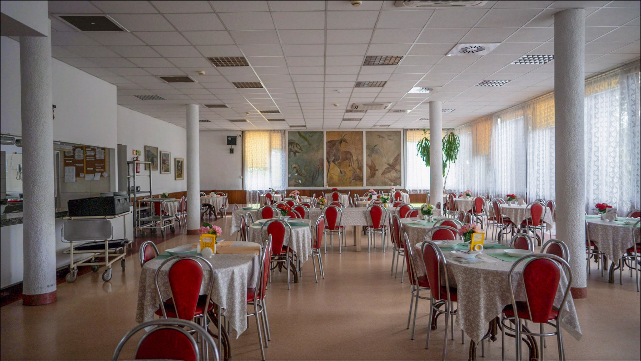 Sala restauracyjna z nakrytymi stołami, czerwone krzesła, białe obrusy w kwiaty, na ścianie tryptyk z motywami zwierzęcymi.