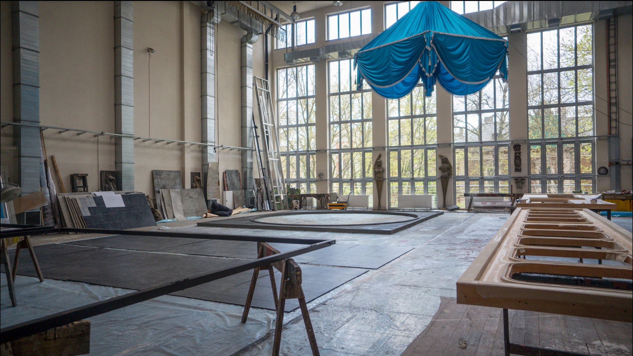 Przestronne wnętrze hali produkcyjnej z dużymi oknami, widoczne elementy scenografii, drewniane konstrukcje i niebieska draperia pod sufitem.