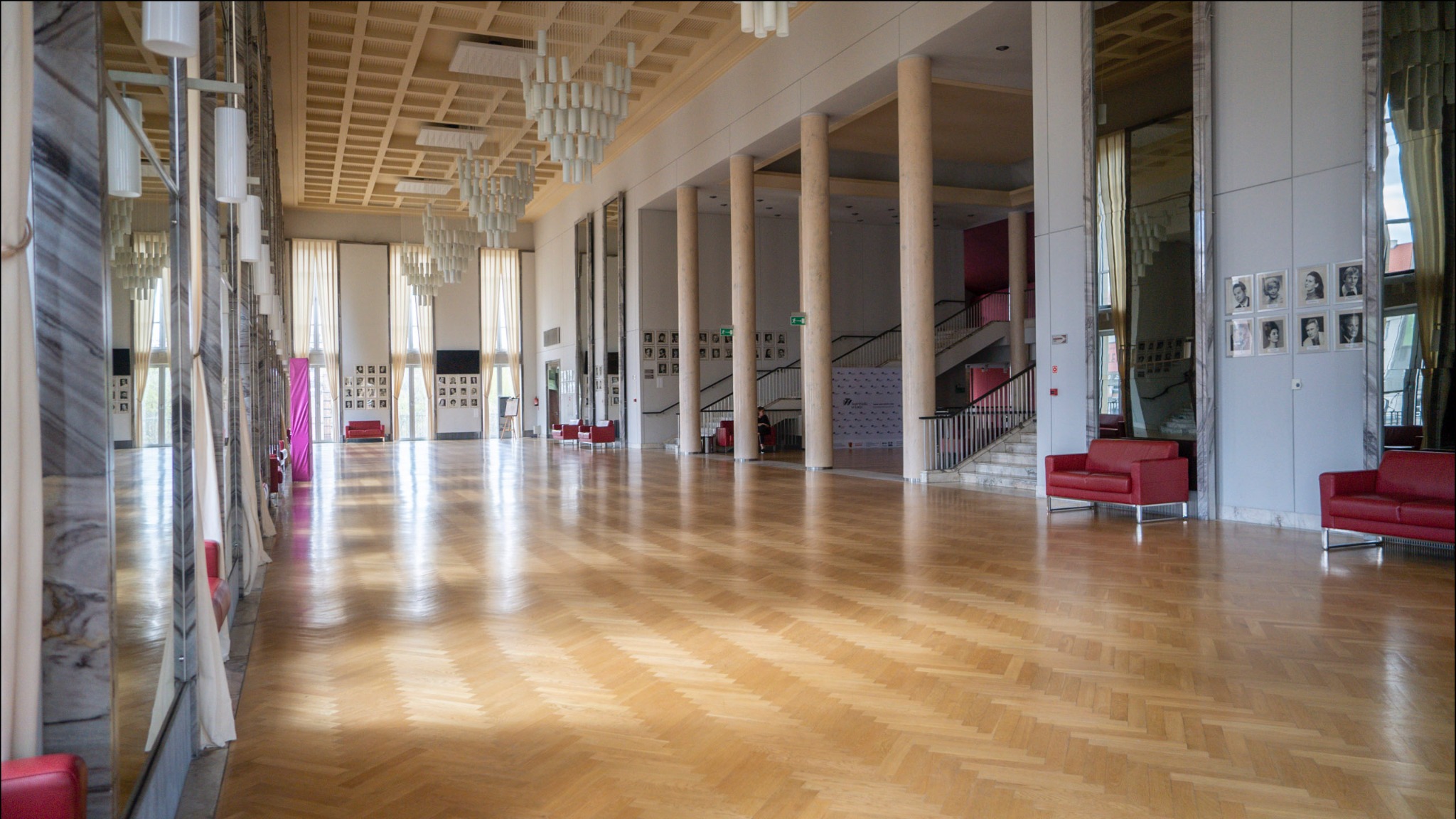 Przestronne wnętrze hali z parkietem ułożonym we wzór jodełki, wysokie kolumny, lustrzane ściany, rzędy lamp i czerwone sofy.