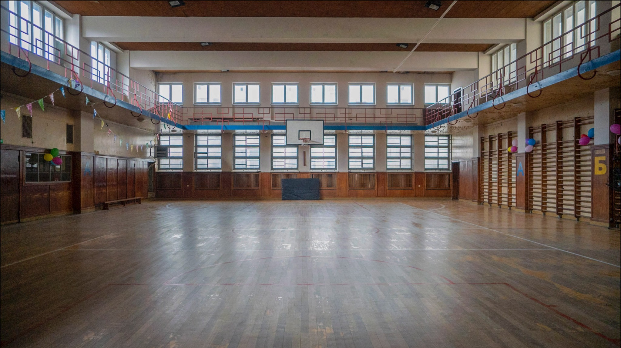 Wnętrze szkolnej sali gimnastycznej z parkietem, drabinkami, oknami i koszem do koszykówki, ozdobionej girlandami i balonami.