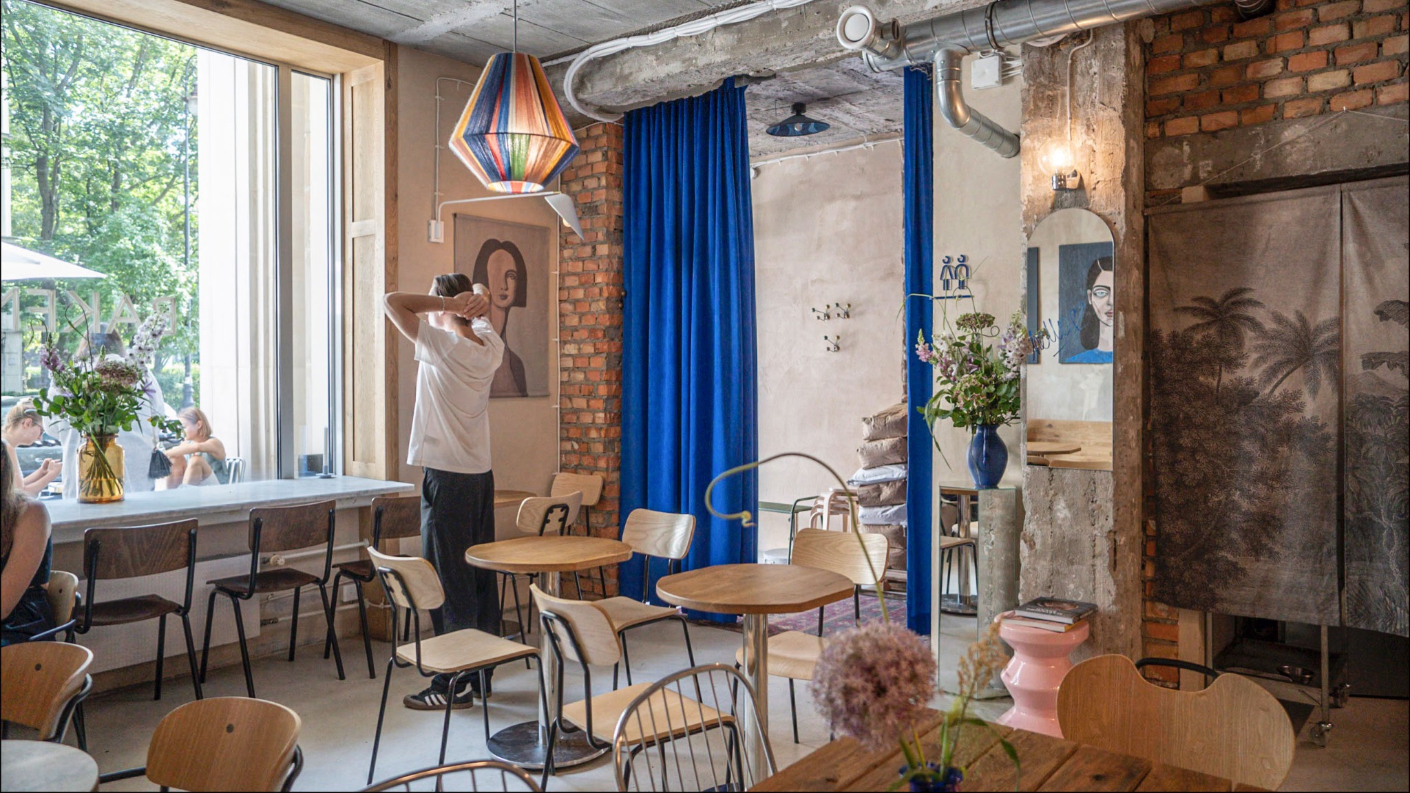 Wnętrze kawiarni z drewnianymi stolikami i krzesłami, dużym oknem z widokiem na zieleń, dekoracyjną lampą wiszącą i elementami cegły na ścianach.