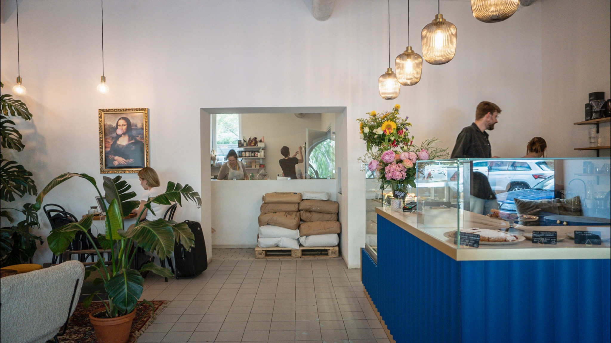 Wnętrze kawiarni z klientką pracującą przy stoliku, dekoracjami roślinnymi, obrazem Mony Lisy z gumą balonową i ladą z ekspozycją ciast.