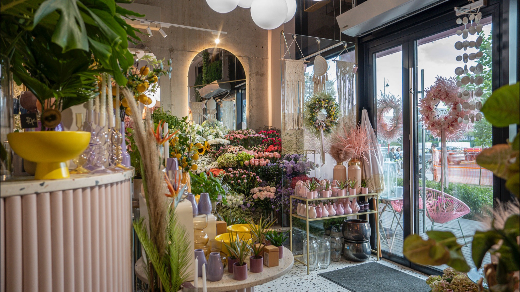 Wnętrze kwiaciarni z bogatą ekspozycją kwiatów, roślin doniczkowych i dekoracji, w tym wianków i makram, widoczne okrągłe lustro i lampy sufitowe.