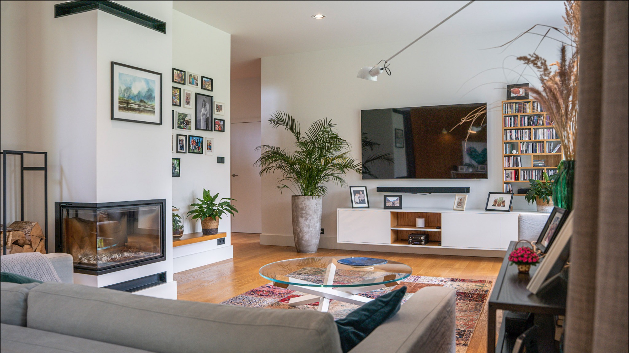 Jasny salon z kominkiem narożnym, drewnianą podłogą, roślinami doniczkowymi i kolekcją zdjęć na ścianie, oświetlony światłem dziennym.