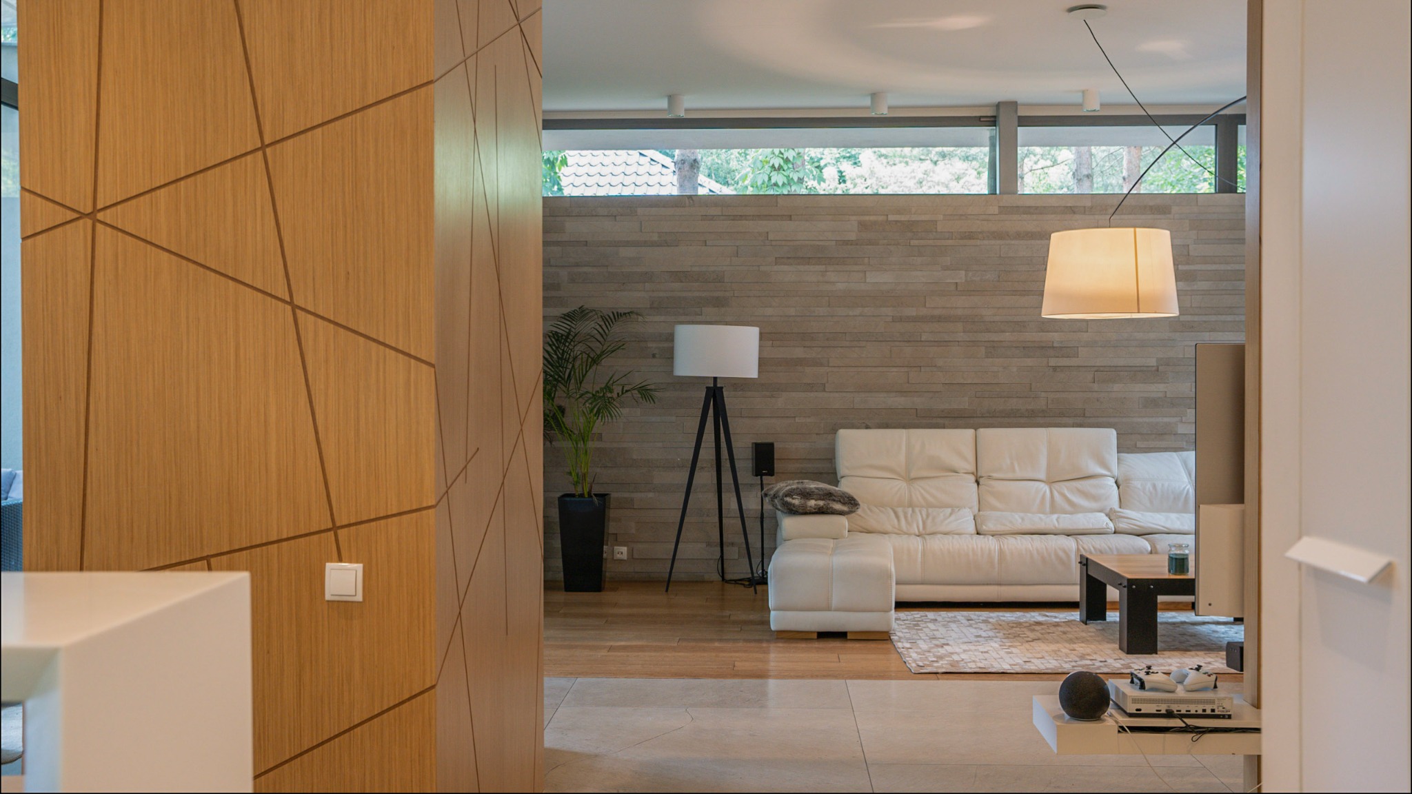 Salon z białym skórzanym narożnikiem, drewnianym stolikiem kawowym i lampą wiszącą, widok zza drewnianej ściany z geometrycznym wzorem.