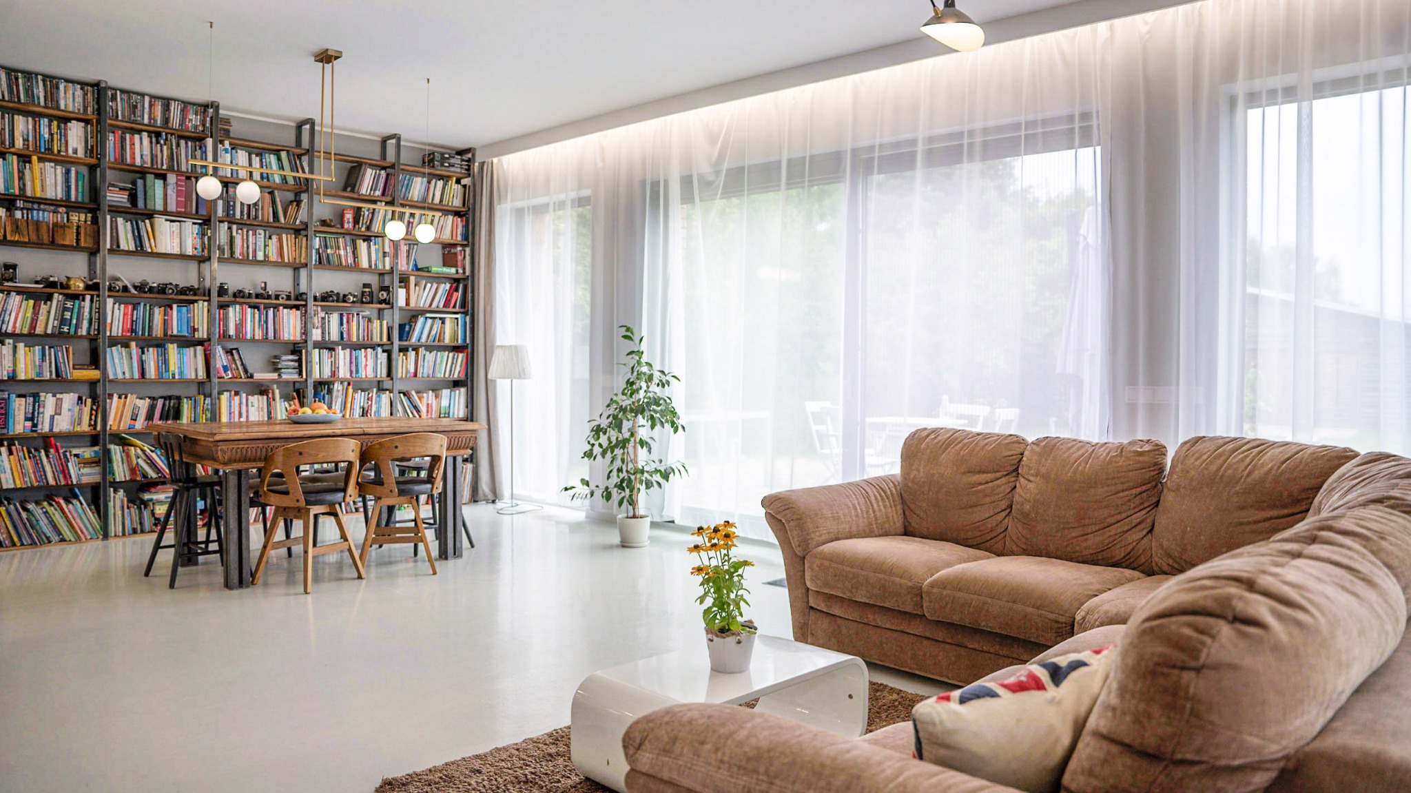 Jasne, przestronne wnętrze z dużą biblioteczką, drewnianym stołem z krzesłami, brązową sofą i białym stolikiem kawowym z kwiatami, oświetlone naturalnym światłem przez duże okna z białymi zasłonami.