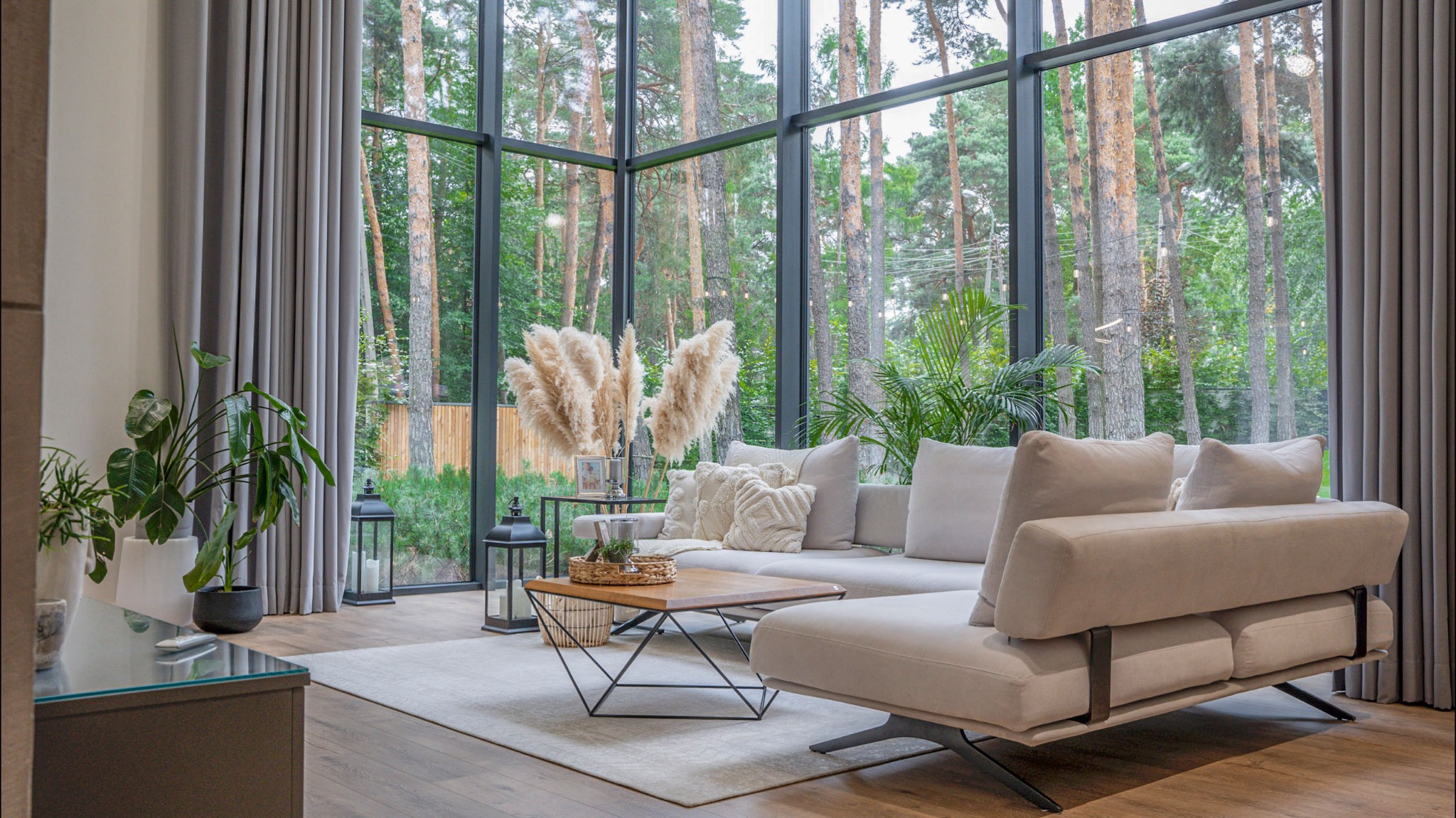 Jasny salon z panoramicznym oknem na las, beżowa sofa narożna z dekoracyjnymi poduszkami, drewniany stolik kawowy z metalową podstawą, wysokie pióropusze w wazonie, rośliny doniczkowe i szare zasłony.