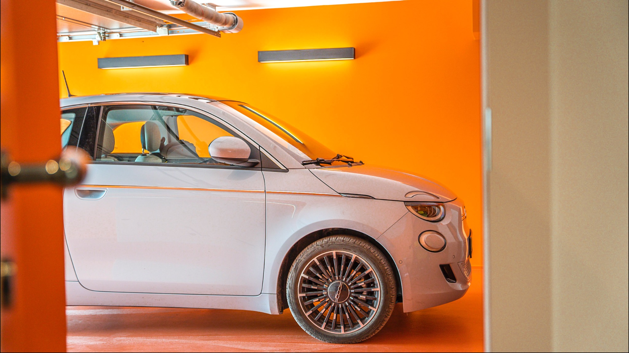 Nowoczesny, jasnoszary samochód elektryczny w garażu z pomarańczowymi ścianami i oświetleniem LED, widok z boku przez częściowo otwarte drzwi.