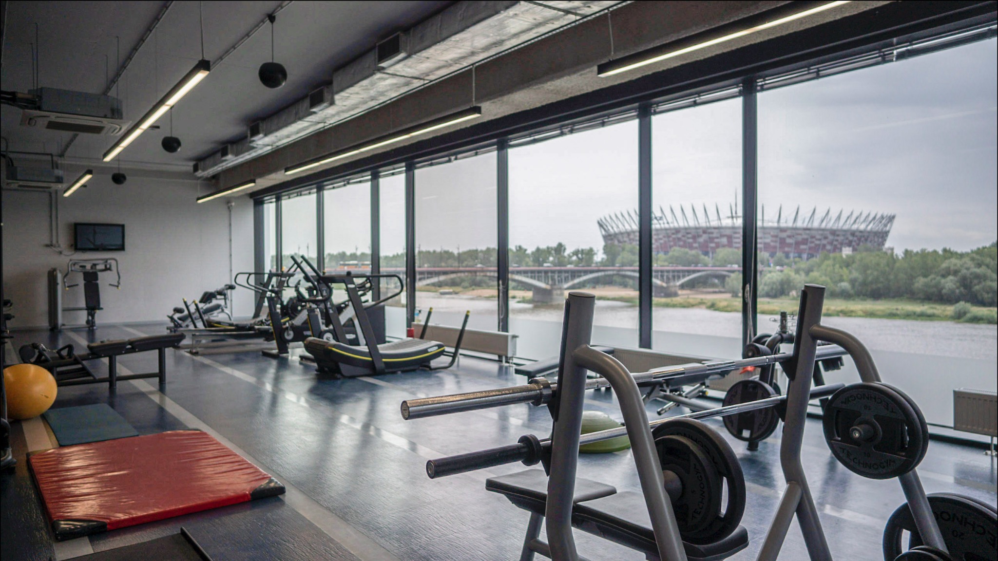 Jasne wnętrze siłowni z widokiem na Stadion Narodowy w Warszawie przez panoramiczne okna, sprzęt do ćwiczeń na pierwszym planie.