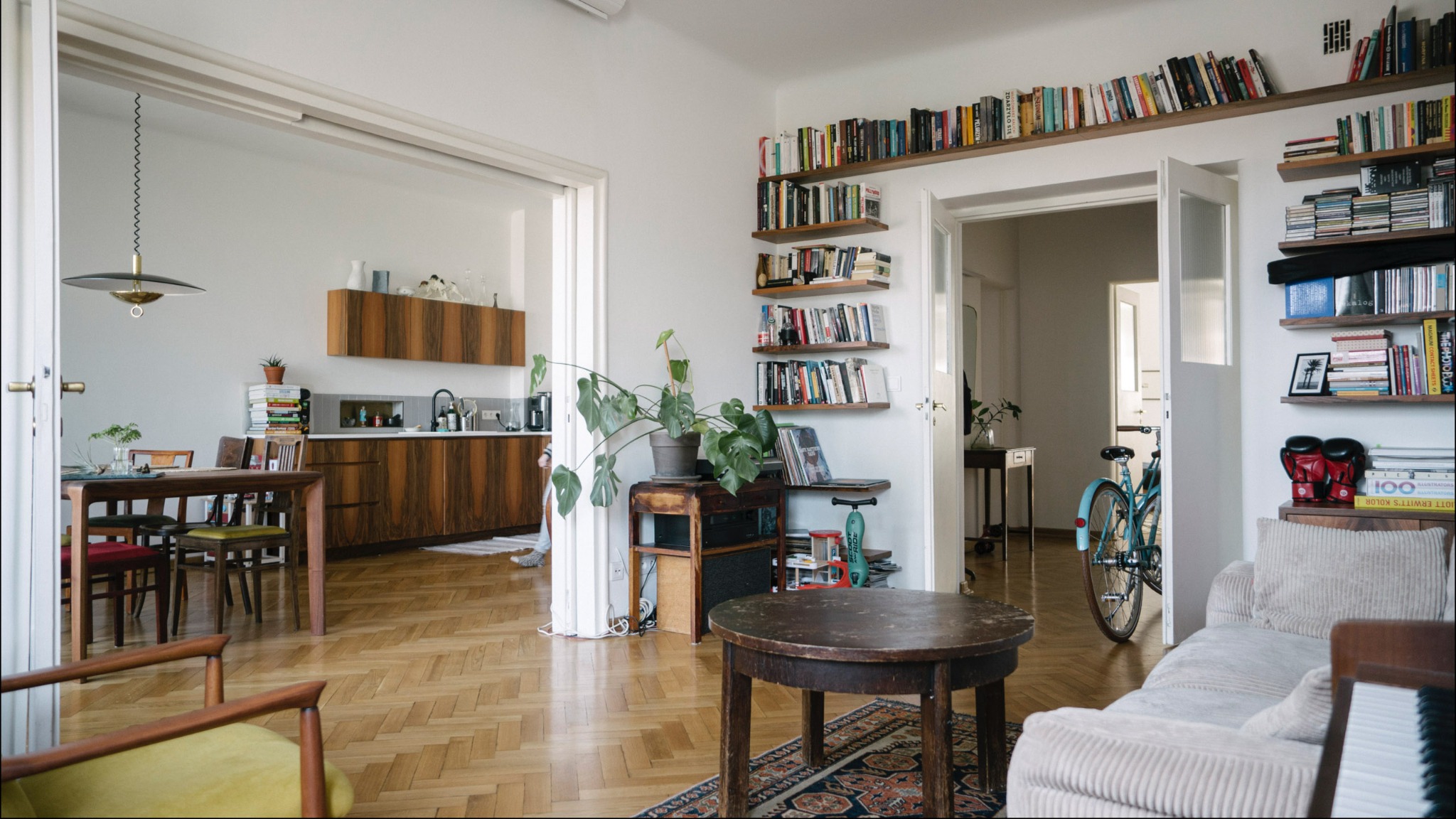Jasne, przestronne wnętrze mieszkania z drewnianą podłogą w jodełkę, widok na kuchnię z zabudową w orzechowym fornirze, regały z książkami na ścianach, okrągły stolik kawowy i rower w tle.