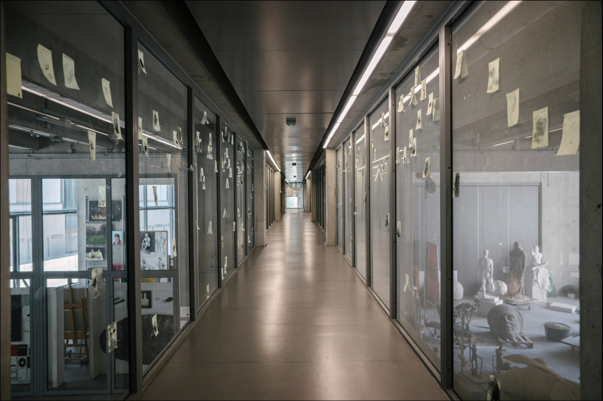 Długi korytarz w pracowni artystycznej, z przeszklonymi boksami wypełnionymi szkicami i rzeźbami, oświetlony liniowymi lampami.