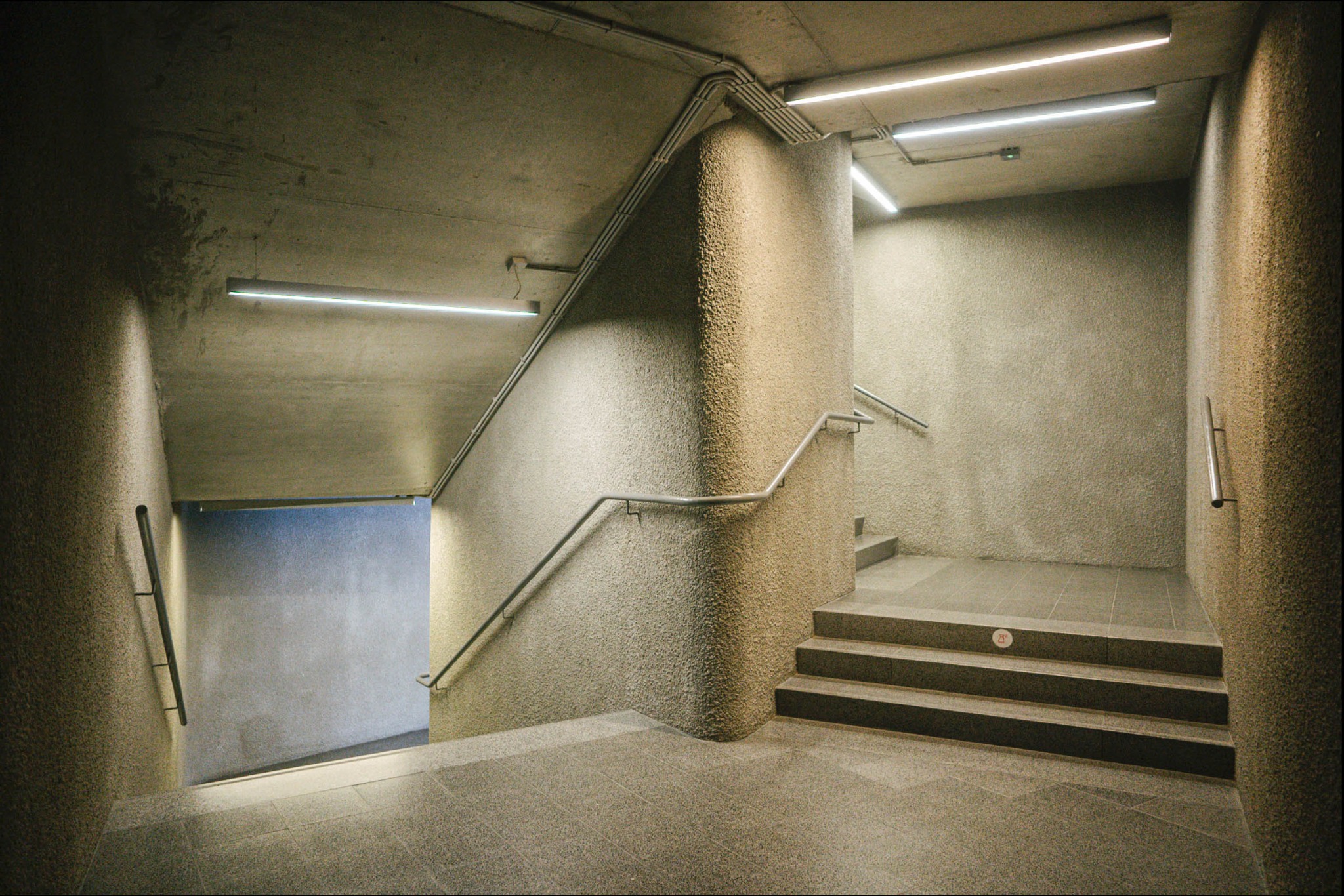 Betonowe schody w dół z metalową poręczą i oświetleniem LED, widoczne rury na suficie.