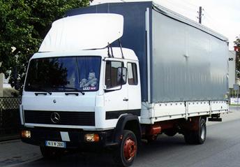 Biała ciężarówka Mercedes z szarą plandeką, zaparkowana na ulicy, widoczny numer rejestracyjny i drzewo w tle.