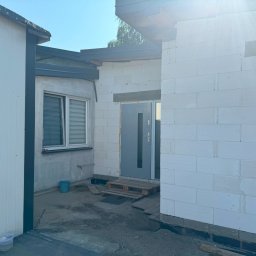 ELTAR Konrad Lamprecht - Budynek w trakcie budowy z białych bloków, widoczna szara stolarka okienna i drzwi wejściowe, paleta przed wejściem, fragment ocieplonego budynku z boku.