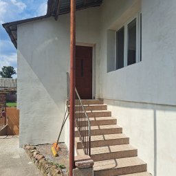 ELTAR Konrad Lamprecht - Wejście do domu po remoncie: otynkowana elewacja, schody wyłożone płytkami, metalowa barierka, oparta o ścianę miotła z żółtym włosiem.