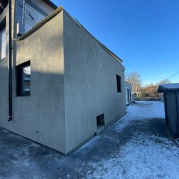 ELTAR Konrad Lamprecht - Szara, otynkowana ściana budynku z ciemnymi oknami i rurą spustową, widoczna w zimowej scenerii z częściowo ośnieżonym brukowanym chodnikiem.