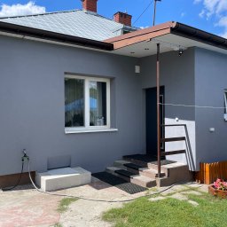ELTAR Konrad Lamprecht - Szary dom jednorodzinny z brązowymi rynnami i schodami wejściowymi. Świeżo pomalowana elewacja w słoneczny dzień, widoczne kwiaty w donicach i fragment ogrodzenia.