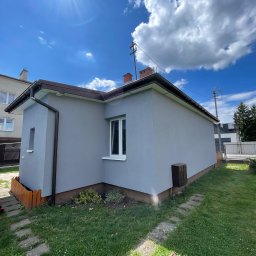 ELTAR Konrad Lamprecht - Szary dom jednorodzinny po renowacji elewacji, widok z boku na tle błękitnego nieba z chmurami. Widoczne detale: brązowe rynny, białe okna, drewniane ogrodzenie.