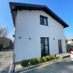 ELTAR Konrad Lamprecht - Biały, dwukondygnacyjny dom z numerem 67, czarną stolarką i dachem, ocieplony, z równo przyciętymi krzewami i szarym brukiem na podjeździe, w słoneczny dzień.