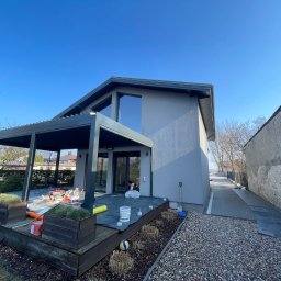 ELTAR Konrad Lamprecht - Elewacja domu jednorodzinnego po malowaniu na szaro, z widocznym tarasem i pergolą. Prace wykończeniowe w toku, narzędzia i materiały budowlane na tarasie.